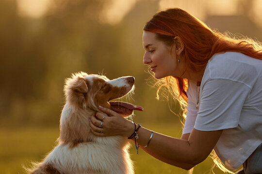 Close-up profile of a happy redhead woman bonding with her Australian Shepherd dog in golden hour sunlight at the park. Concept of love, pet care, animal friendship and trust in nature 