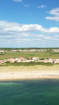 Scenic aerial shot of a sandy beach, clear sea, and traditional village rooftops on Ile de Re under a blue sky with clouds.