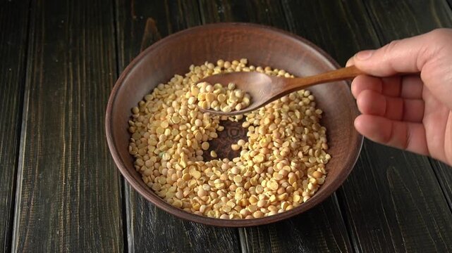 Hand scooping dried yellow peas with wooden spoon from a brown bowl on a rustic wooden table, showcasing the action of serving ingredients