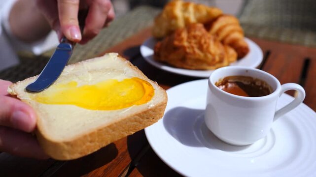 Close up of a person's hands spreading delicious orange jam on a slice of buttered bread during a sunny breakfast, with coffee and croissants served on a wooden table in the background