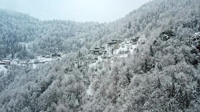 Aerial view of snow-covered forest and mountain houses during snowfall in the Ayder area of Rize, Turkey. Winter landscape with wooden homes and white trees creates a peaceful mountain scene.