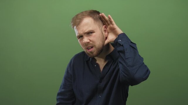 Man cups ear and leans forward in green studio set, listening for a faint sound from offstage while squinting and tilting head; curiosity.