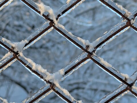 Metal wire mesh fence covered in thick ice and snow. Geometric pattern of frozen chain link fence after freezing rain. Winter background with icy grid and snow. Glazed frost on wire.