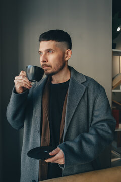A man in a gray coat with a black mug of coffee against the wall in a cafe interior in spring.