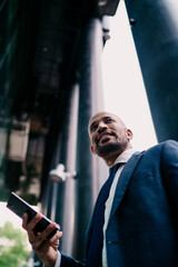 Vertical shot of smiling businessman holding smartphone against tall office columns, portraying...