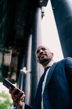 Vertical shot of smiling businessman holding smartphone against tall office columns, portraying success, digital freedom and confidence in flexible, tech-driven freelance career.