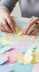 Image shows hands arranging a yellow paper butterfly. Colorful craft paper and floral pattern beneath symbolize crafting, creativity, and artistry