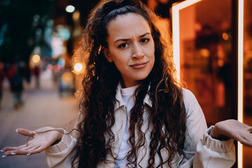 Curly-haired woman shrugs with puzzled face in city street, showing questioning emotion and digital miscommunication in daily mobile experience. © BullRun