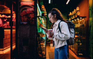 Fototapeta premium Young woman walks past colorful storefronts, holding smartphone and coffee, immersed in digital routine while navigating urban evening filled with artificial light.