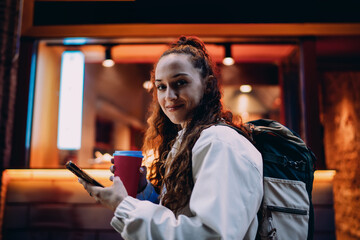 Smiling young woman stands near food kiosk with backpack, holding smartphone and coffee, combining casual fashion with tech-enabled lifestyle in city night glow. © BullRun