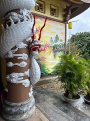 White dragon statue with red accents resting on a decorative brown base, set against a vibrant mural of a serene landscape and lush green palm trees. © Karina