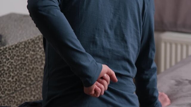 A man sitting on a bed rubs or massages his back for pain in the blue. Concept of back pain