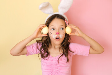 A girl with mouth open in surprise, shows yellow Easter eggs with a painted bunny face.