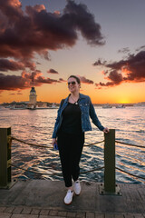 Fototapeta premium Mujer feliz al atardecer con la icónica Torre de la Doncella (Kız Kulesi) de fondo en Estambul. Cielo dramático y nubes coloridas sobre el Bósforo durante la hora dorada.