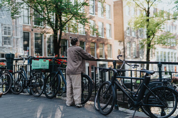 A woman stands by parked bicycles on a canal bridge in Amsterdam, Netherlands. The scene shows a quiet urban canal with bikes and historic buildings lining the water.