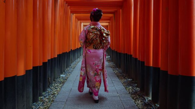 Asian woman wearing traditional kimono walking through the famous red torii gates at Fushimi Inari Shrine in Kyoto, Japan. Cultural travel destination showcasing historic Japanese heritage and Shinto 