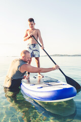 Photo of cute little surfer and his dad in the sea. Photo of a young boy trying out to hold a balance on a paddle board for the first time. His father are beside him, giving him support.