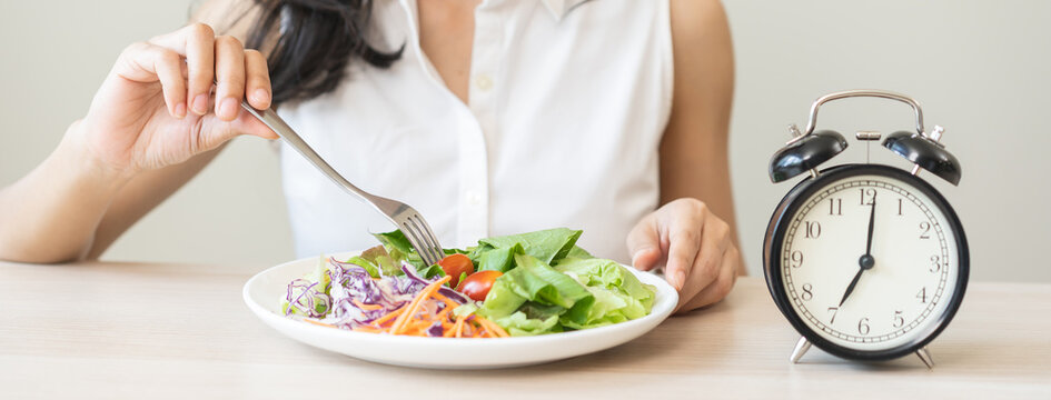 Intermittent fasting with clock, health asian young woman, girl weight loss, eating green fresh vegetable salad on dish, plate with eat healthy of breakfast food in morning, lunch on a table at home.
