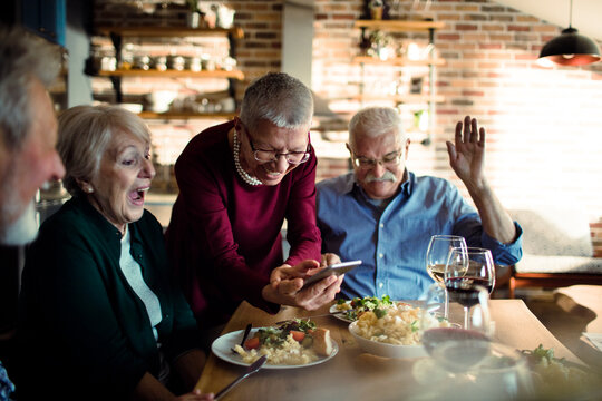 Senior friends share a laugh over smartphone during dinner at home
