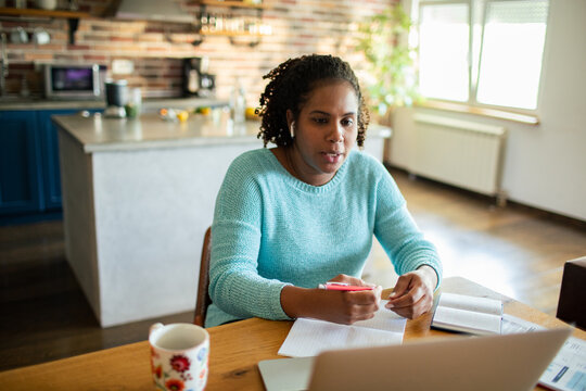 Woman taking notes on video call in home kitchen
