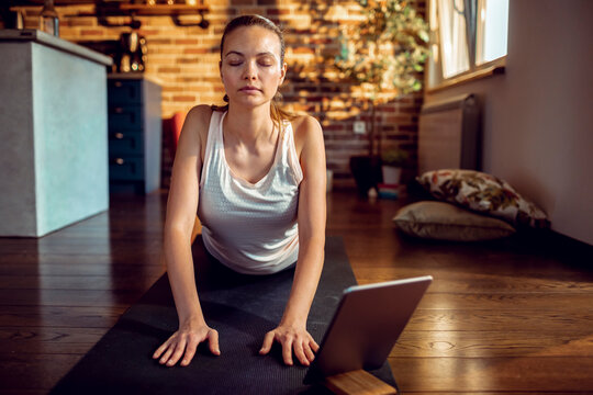 Young woman practicing yoga cobra pose at home with tablet