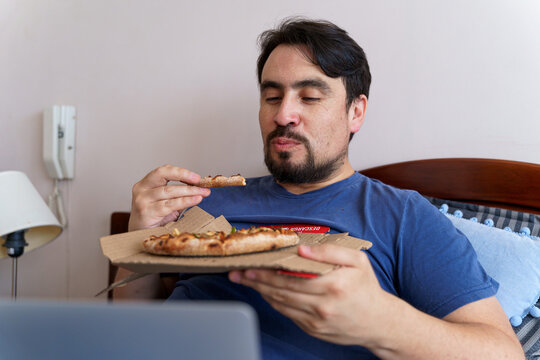 A tight portrait of a man taking a satisfying bite of pizza while keeping his eyes fixed on an off-camera screen. The image perfectly captures the engagement of binge-watching during a casual home mea