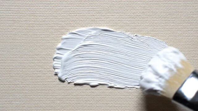 A paintbrush spreads white acrylic paint on a textured beige surface, demonstrating brush strokes from initial to full coverage with thick, layered paint.