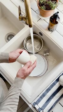 Man washes dishes in a kitchen sink with a sponge and liquid soap under running water. Contemporary domestic scene and modern masculinity. Concept of sharing household chores. Day light, close-up
