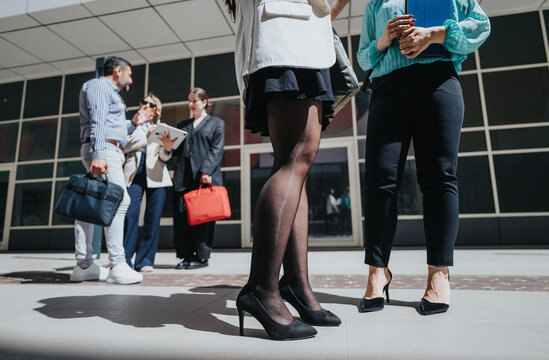 Business colleagues stand outside a modern office building engaged in conversation. A small group of professionals gathers near the entrance holding bags and documents.