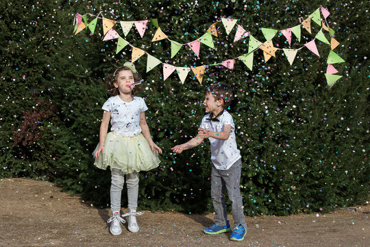 Happy Boy and girl standing side by side in a garden throwing confetti in the air and blowing a party blower at a summer party