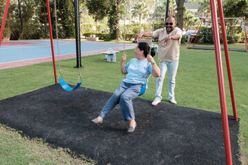Man pushes woman on swing in park while enjoying sunny day outdoors with green grass and trees