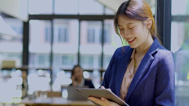  A cheerful businesswoman stands in her office, using a digital tablet to work on a financial project.