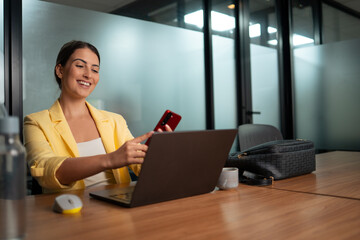 Young professional woman in yellow blazer using smartphone while working on laptop in modern corporate office workspace
