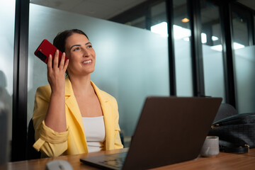 Professional woman in yellow blazer listening to voice message on smartphone in modern corporate office workspace