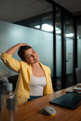Professional woman in yellow blazer doing neck stretch exercise during work break at desk in modern corporate office