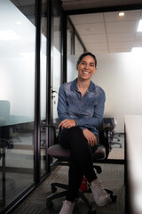 Portrait of smiling professional woman in denim shirt sitting on office chair in modern corporate workplace