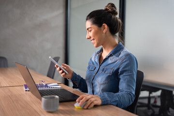 Smiling professional woman in denim shirt checking smartphone while working on laptop in modern corporate office