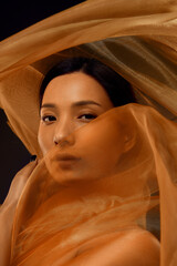 Fototapeta premium Asian woman cinematic portrait with face partially hidden behind translucent golden silk fabric. Luxury aesthetic for cosmetics advertising, beauty branding, fashion editorials and skincare campaigns.