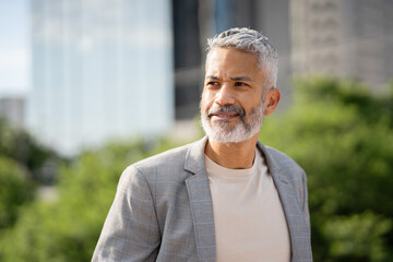 Mature professional man with gray beard in blazer standing outdoors with modern buildings and green trees in urban background