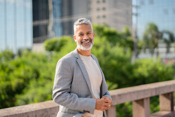 Naklejka premium Mature Brazilian businessman with gray beard in blazer standing outdoors in modern urban city with green landscape
