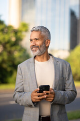 Smiling mature Brazilian businessman with gray beard holding smartphone outdoors in modern urban city environment