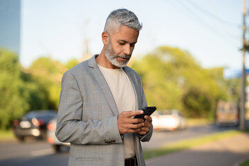 Focused mature Brazilian businessman with gray beard using smartphone on urban sidewalk with cars and green trees