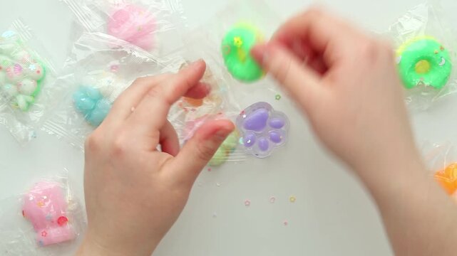 Top view of hands on a white table opening a clear wrapper, picking up a green donut squishy, pressing and squeezing it repeatedly, then holding the soft toy in a playful sensory moment.