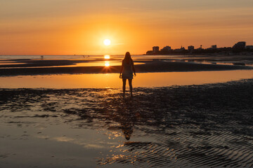 Woman standing on a driftwood log on wide tidal flats with ripple patterns in the sand at low tide. Minimalist coastal landscape with calm sea and setting sun in the background.