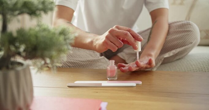 Woman painting her fingernails with pink nail polish at home in a cozy interior