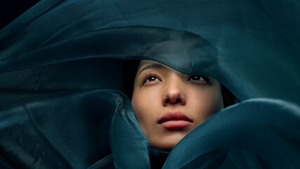 Cinematic beauty portrait of Asian woman emerging through flowing teal silk fabric, luxury fashion...