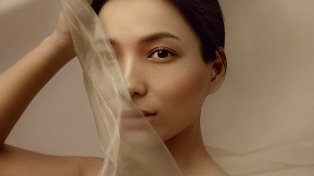 Elegant beauty portrait of Korean woman partially covered by translucent beige silk fabric symbolizing softness and modern luxury aesthetics for cosmetics advertising and beauty campaigns.