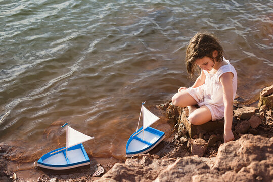Side view of a young Girl sitting on rocks by a lake playing with two toy boats, Linares, Provincia de Jaen, Andalucia, Spain