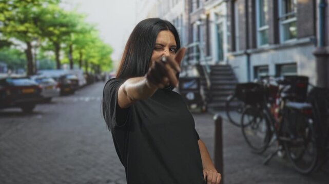 Woman pointing finger at camera on street while winking and smiling, wearing black tshirt and standing before bicycles and row buildings; confidence.