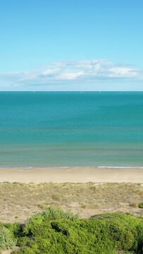Wide view of a sandy beach with turquoise water and clear blue sky at Isola Varano Nature Reserve, Italy. Lush greenery and natural dunes frame the tranquil coastal landscape.